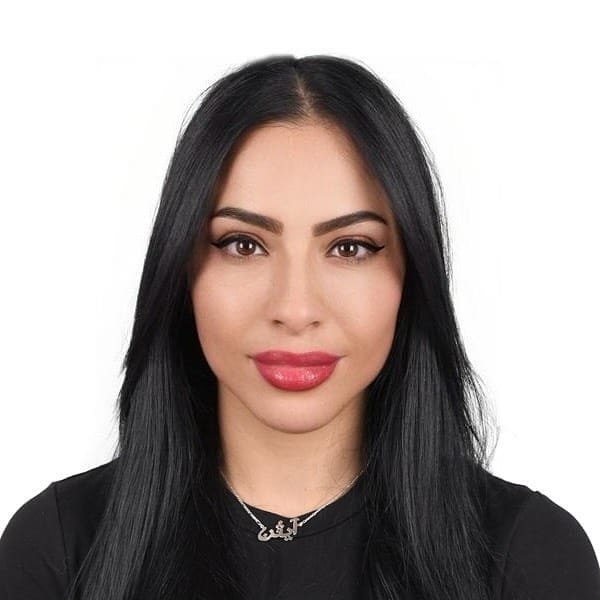A person with long, straight black hair and smooth skin is wearing a black top and a necklace with small pendants. They have prominent eye makeup, including winged eyeliner, and glossy pink lipstick. The background is plain white.